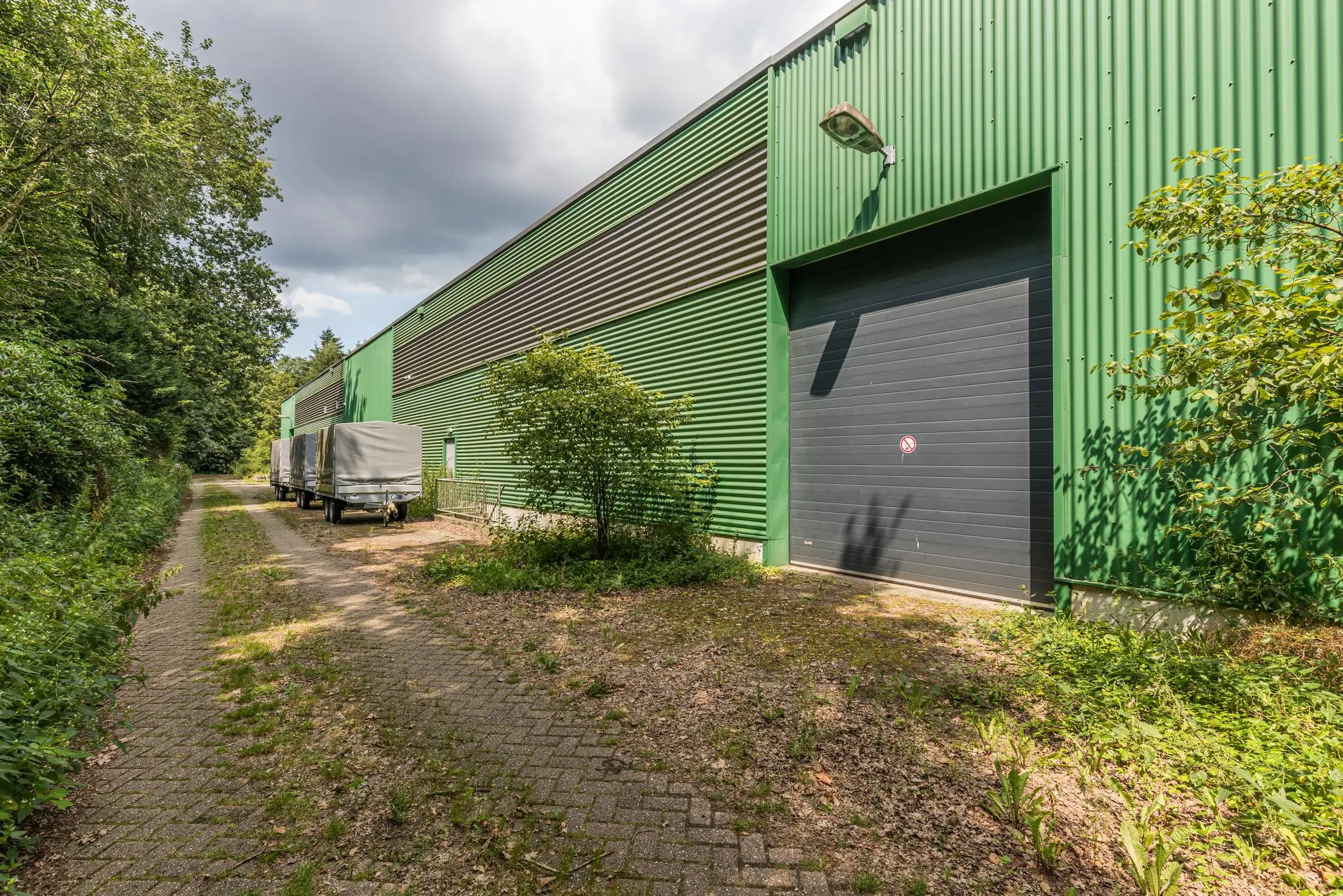 Groene loods met grote garagedeur en geparkeerde aanhangers aan de Laan Blussé van Oud-Alblas, omringd door bomen en groen.